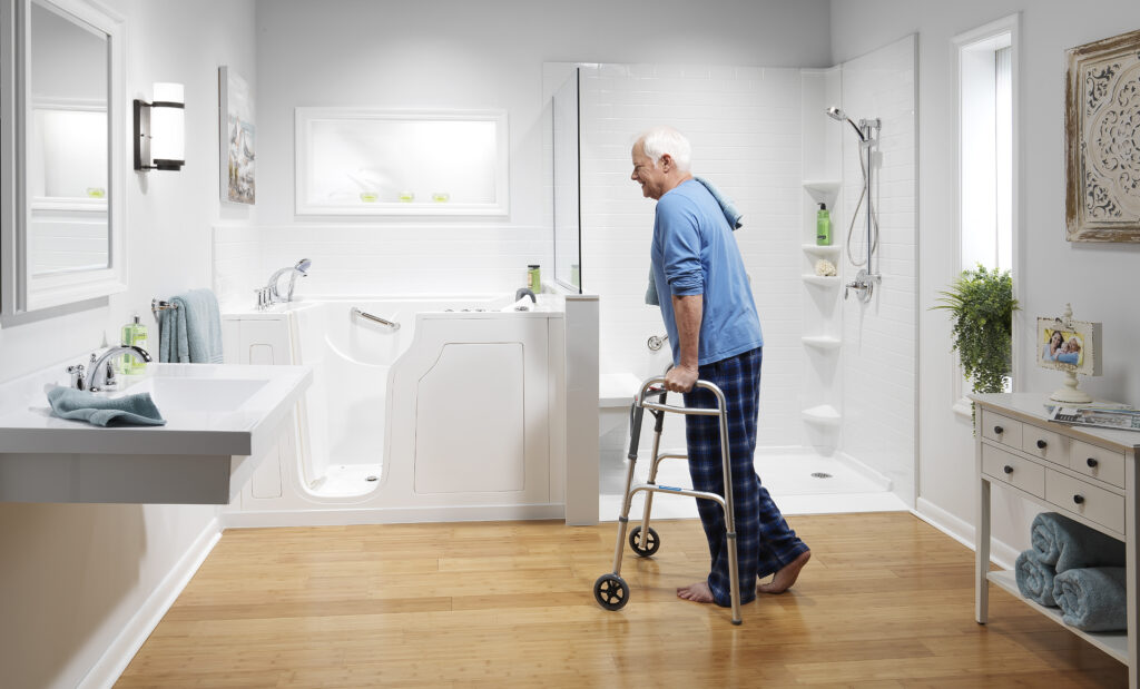 Older man using a walker in a bright, modern bathroom with a glass shower and white tiled walls.