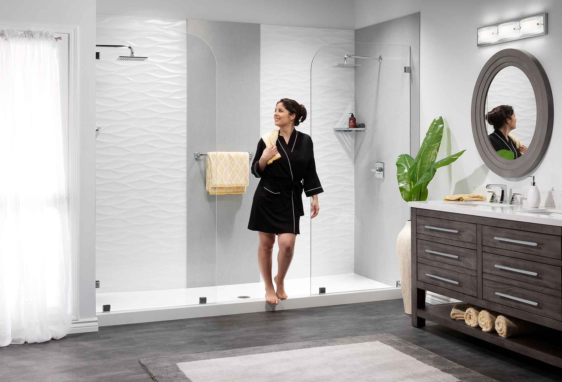 Woman in a black bathrobe standing inside a glass-walled shower in a modern bathroom with a round mirror nearby.