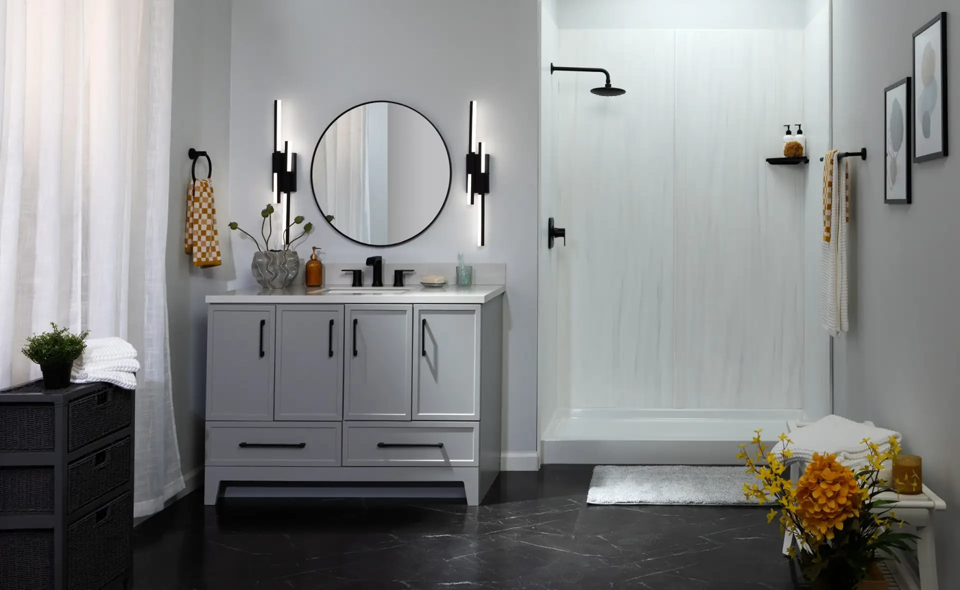 Modern gray bathroom with a round black-framed mirror above a gray vanity, vertical black light sconces, and a glass-enclosed shower with a rainfall head; yellow towels and flowers as accents.