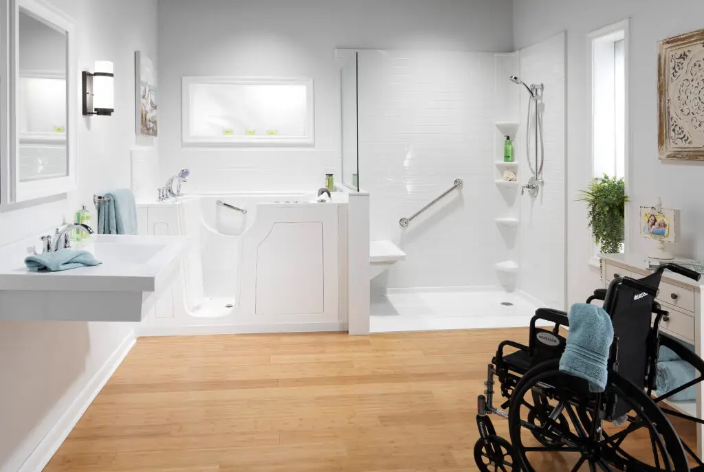 Wheelchair-accessible bathroom remodel showcasing a white walk-in safety tub adjacent to a barrier-free shower with an angled grab bar, subway tile walls, and an empty black wheelchair in the foreground.