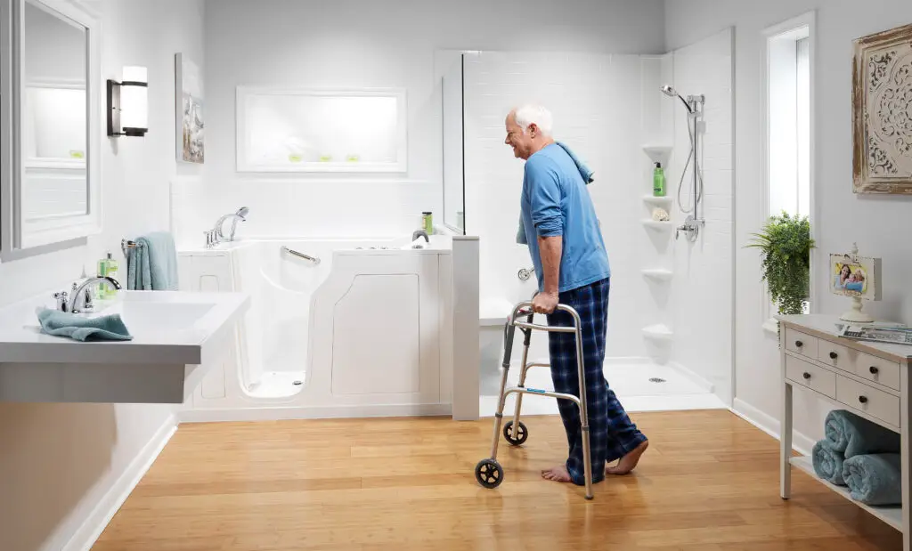 An elderly man using a walker navigates a spacious handicap-accessible bathroom featuring a matching white walk-in safety tub and a low-barrier tile shower combo.