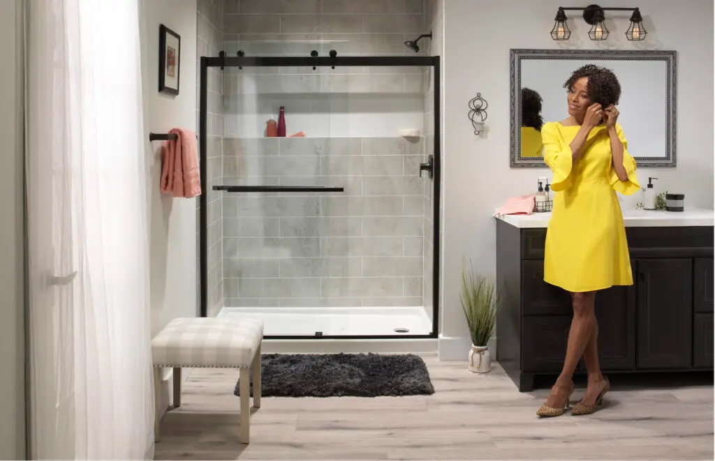 Modern bathroom remodel featuring a walk-in shower with weathered grey concrete-look tile walls, a matte black rolling barn-style glass door, a dark vanity, and a woman in a yellow dress.