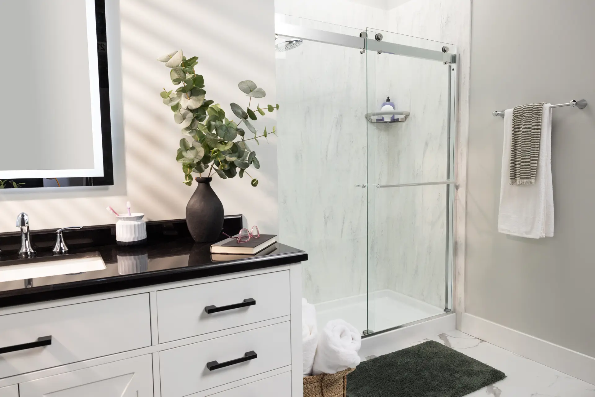 Modern bathroom remodel showing a walk-in shower with a frameless sliding glass door, textured light grey stone walls, and a white vanity with a black countertop.