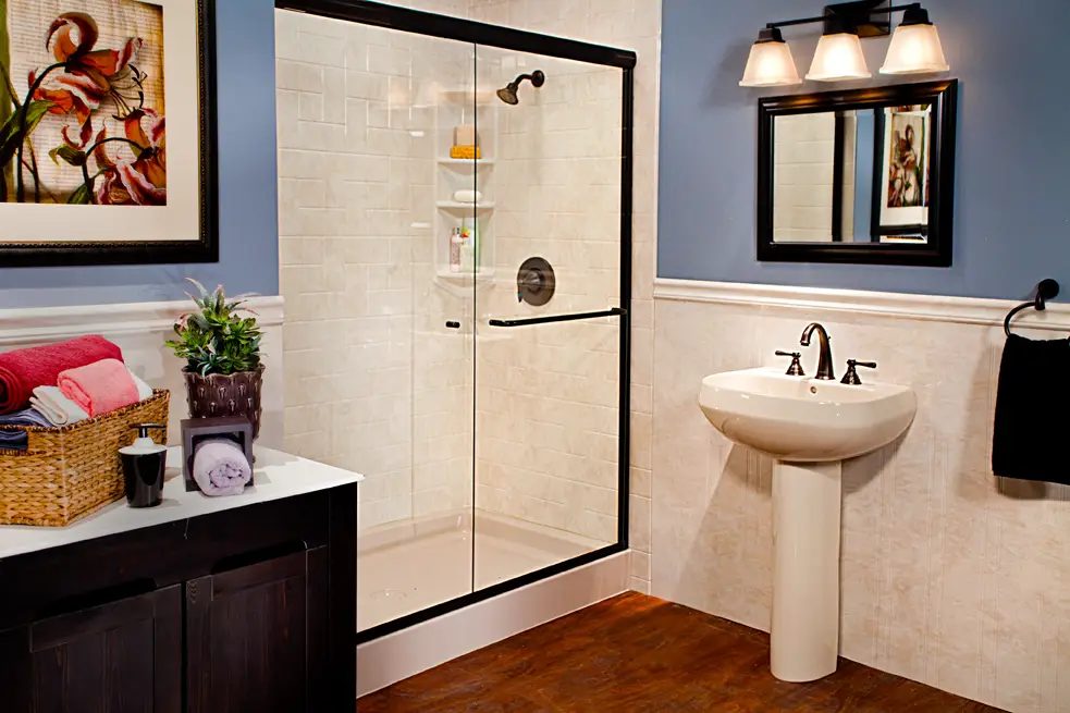 Traditional bathroom remodel featuring a walk-in shower with travertine windmill tile walls, a matte black frame sliding door, a white pedestal sink, and matching stone look wainscoting panels against blue walls.
